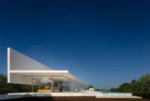 A modern white glass house with a flat roof and large windows overlooking a clear blue sky and greenery.