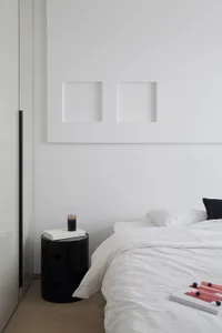 A modern minimalist bedroom with a textured white blanket on a cozy bed, accompanied by a sleek, black side table with a glas