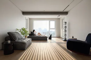 A modern minimalist living room with light wood flooring, a dark lounge chair, a gray sofa, and a woman sitting on a daybed w