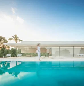 A modern minimalist pool house with a white facade, glass windows, and concrete patio.