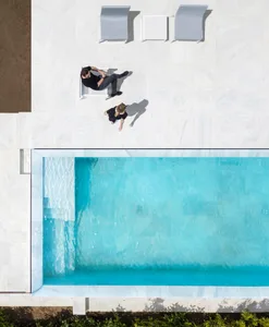 A minimalist outdoor lounge area with marble elements, featuring a white marble patio, chairs, and a swimming pool.