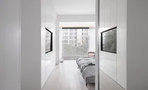 Modern minimalist bedroom with white cabinets and a bed, featuring large windows with a view of urban buildings and greenery.