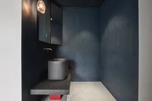 A sleek, minimalist modern bathroom with dark concrete walls, a circular sink, and a red accent.