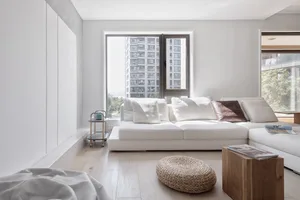 Modern, minimalist living room with light-colored wooden flooring, white furniture, and large windows.