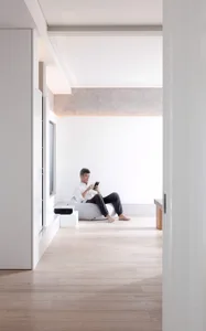 Person sitting on a beige fabric ottoman in a minimalist living room, looking at a smartphone, floor-to-ceiling white walls, 