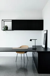 A minimalist workspace featuring a black wooden desk, a beige chair, a black lamp, and a framed black and white abstract pain