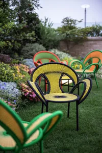 A modern, yellow, geometric-patterned garden chair made of metal and woven fabric surrounded by colorful flowers and plants.
