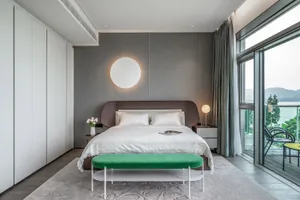 A modern bedroom with white furniture, a green ottoman, and a view of mountains outside.