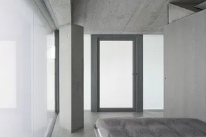 A minimalist bedroom with concrete walls, floor, and ceiling, featuring a bed with a gray cover.