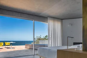 Modern residential bedroom featuring a minimalist design with a light concrete floor, white sheer curtains, and a view of the