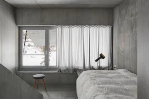 A minimalist bedroom with concrete walls, a white bed, and a white curtain at the window.