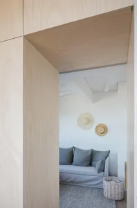 A minimalist living room with a white sofa, grey cushions, and a light wooden wall and floor.