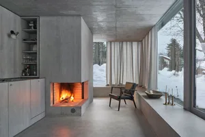 A modern living room with a concrete fireplace, minimalist decor, and large windows offering a snowy landscape view.