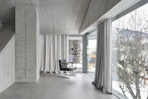A minimalist living room with concrete walls, floor-to-ceiling windows, and a black armchair.