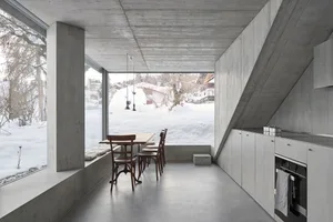 A modern kitchen with concrete flooring, minimalist design, and large windows overlooking a snowy landscape.