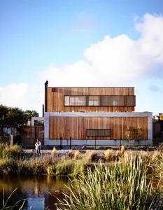 A modern residential home with wooden exterior and large windows, located near a small pond with tall grasses in the foregrou