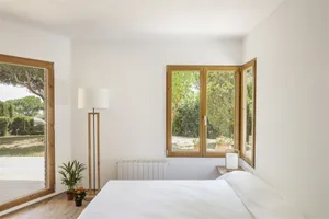 White minimalist bedroom with light wooden furniture and large windows overlooking greenery.