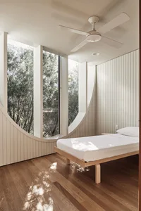 A minimalist bedroom featuring a wooden platform bed in front of a large curved window overlooking trees.