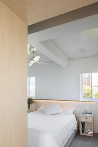 A modern minimalist bedroom featuring a bed with white sheets and a wooden headboard against a white wall, supplemented by am