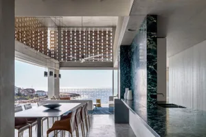 A modern residential kitchen with concrete and marble surfaces, featuring a large table and chairs with a view of the ocean.