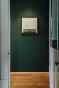 A minimalist interior featuring a framed painting on a dark green paneled wall, with a wooden floor.