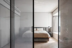 A minimalist bedroom with a modern design, featuring a bed with a cream-colored duvet and a large window letting in natural l