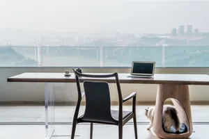 A minimalist workspace featuring a wooden table with a laptop, a black chair, and a cat underneath the table.