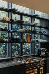 A modern kitchen with metal and glass finishes, featuring a stove, open shelves, and a view of greenery through large windows