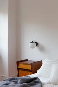 A minimalist bedroom featuring a white wall with a sleek, modern light fixture, a wooden bedside table, and striped bedding.