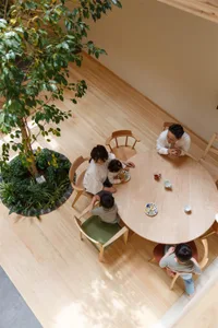 A family sitting at a wooden dining table with green chairs, surrounded by indoor plants and a wooden floor.