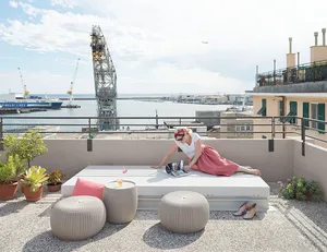 A woman relaxing on a rooftop terrace overlooking the harbor. Concrete floor, minimalistic design, and industrial view.