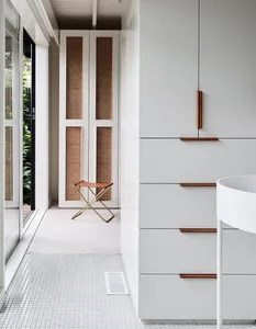 A minimalist bathroom featuring white tile flooring with a foldable brass chair in front of an open closet with cane doors.