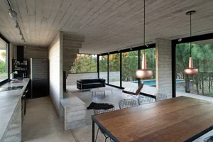 Modern kitchen with concrete walls, dining table, and living room with large windows and outdoor patio.
