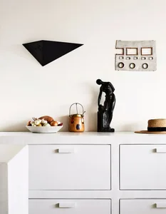 A minimalist kitchen with white cabinets, concrete countertops, a bowl of colorful vegetables, and a black sculpture on the c