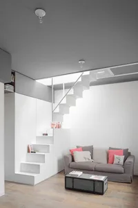 A modern minimalist living room with stairs, concrete flooring, and a gray sofa with pink and white cushions.