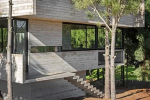 A modern residential exterior made of concrete with large glass windows and a tree in the foreground.