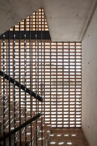 A modern staircase featuring a grid of wooden slats with concrete walls and black metal railings, illuminated by sunlight.