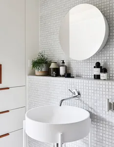 A minimalist modern bathroom with a white ceramic sink, chrome faucet, round wall mirror, and small potted plant on top of a