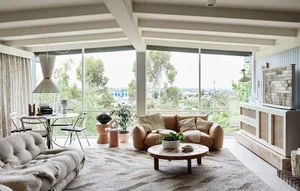 A modern living room with a tan leather sofa, round coffee table, and large windows overlooking greenery.