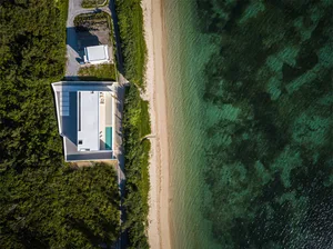 A minimalist beach house with a clay roof, surrounded by greenery and a sandy beach.