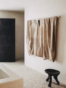 A textured, beige silk wall hanging in a modern bathroom with white pebble tile flooring and a large black framed mirror.