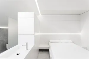 A modern minimalist bedroom with white furniture, a bed with white bedding, and a white sink with a mirror.