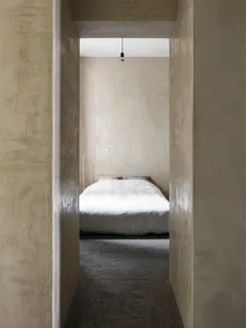 A minimalist bedroom with concrete walls and floor, featuring a white bed and a hanging light bulb.