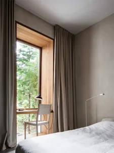 A minimalist bedroom with neutral-colored walls, curtains, and bedding. A small desk with a lamp and chair is placed next to 