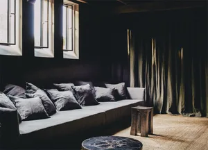 Industrial-style living room with a long gray couch adorned with multiple fabric cushions, large windows with curtains, and a