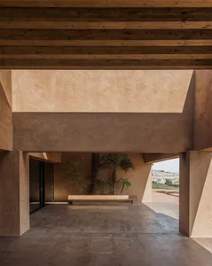 A minimalist indoor design featuring concrete walls and floors, with a central seating area and palm trees in the background.