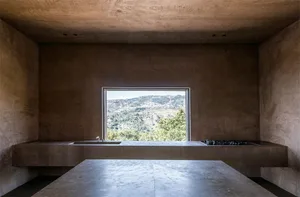 A minimalist kitchen with a concrete countertop and a large window overlooking mountains and trees.
