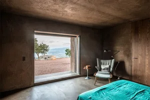A modern bedroom with a turquoise bedspread, a concrete floor, and a large window overlooking a scenic landscape.