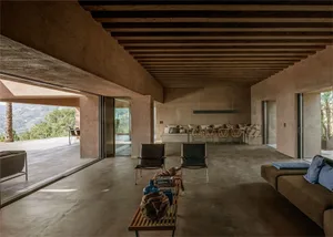 A modern living room with concrete flooring, minimalist furniture, and large windows providing a view of the outdoor area.