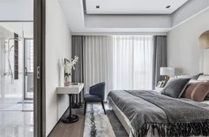 A minimalist bedroom with white curtains, a bed with a gray and brown blanket, and a small table with a vase of flowers and a